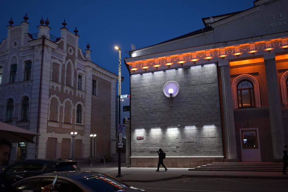 People walking by illuminated historic buildings on a city street at night.